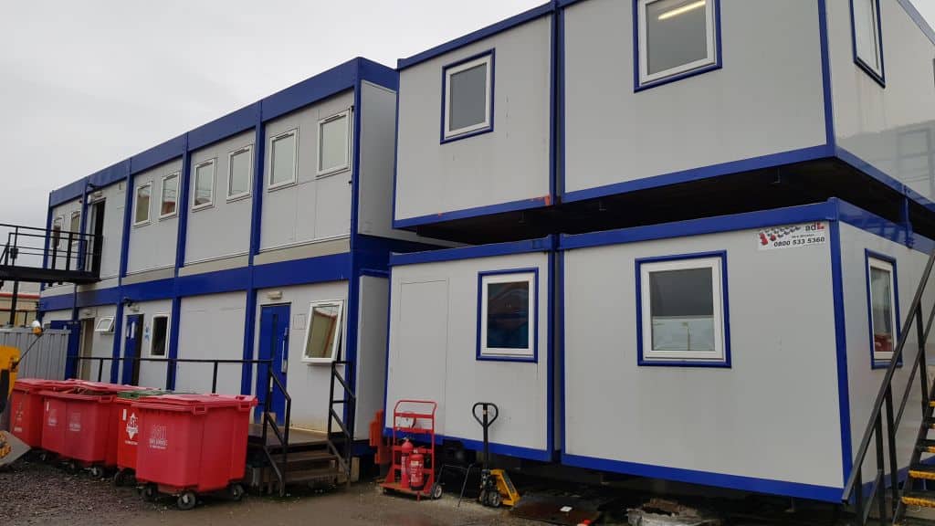 A two-story temporary modular building, featuring blue and white units with windows and a stairway, surrounded by a gravel lot with red garbage bins and a hand pallet truck. Text on sign: 