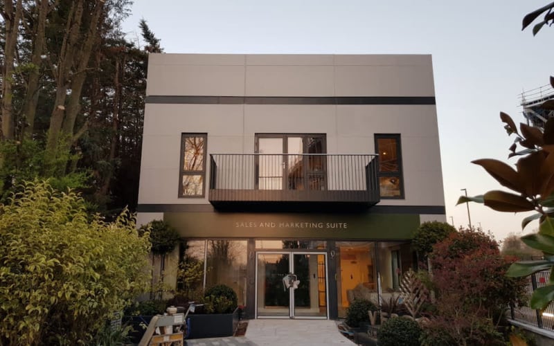 A modern building with labeled entrance, "SALES AND MARKETING SUITE," framed by lush greenery and tall trees. It features a central balcony and large windows, set in a landscaped pathway.