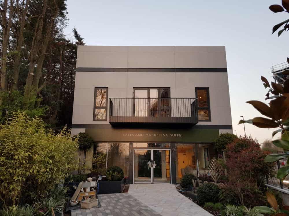 A modern building with labeled entrance, "SALES AND MARKETING SUITE," framed by lush greenery and tall trees. It features a central balcony and large windows, set in a landscaped pathway.