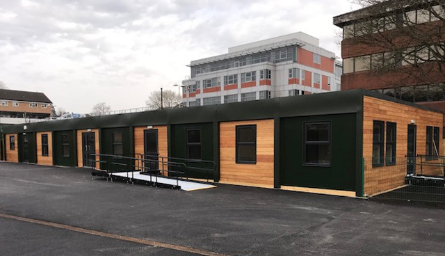 A long, single-story modular building with wooden and dark green panels stands in a parking lot. Metal ramps and stairs provide access. Multi-story buildings and marked parking spaces surround it.