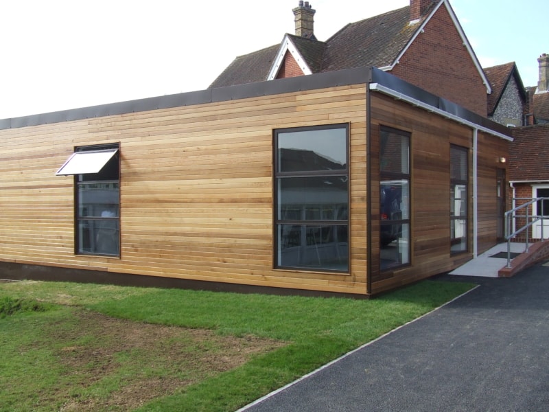 A modern wooden building features large windows, one with an open awning, situated on a grassy lawn with an adjoining concrete walkway.