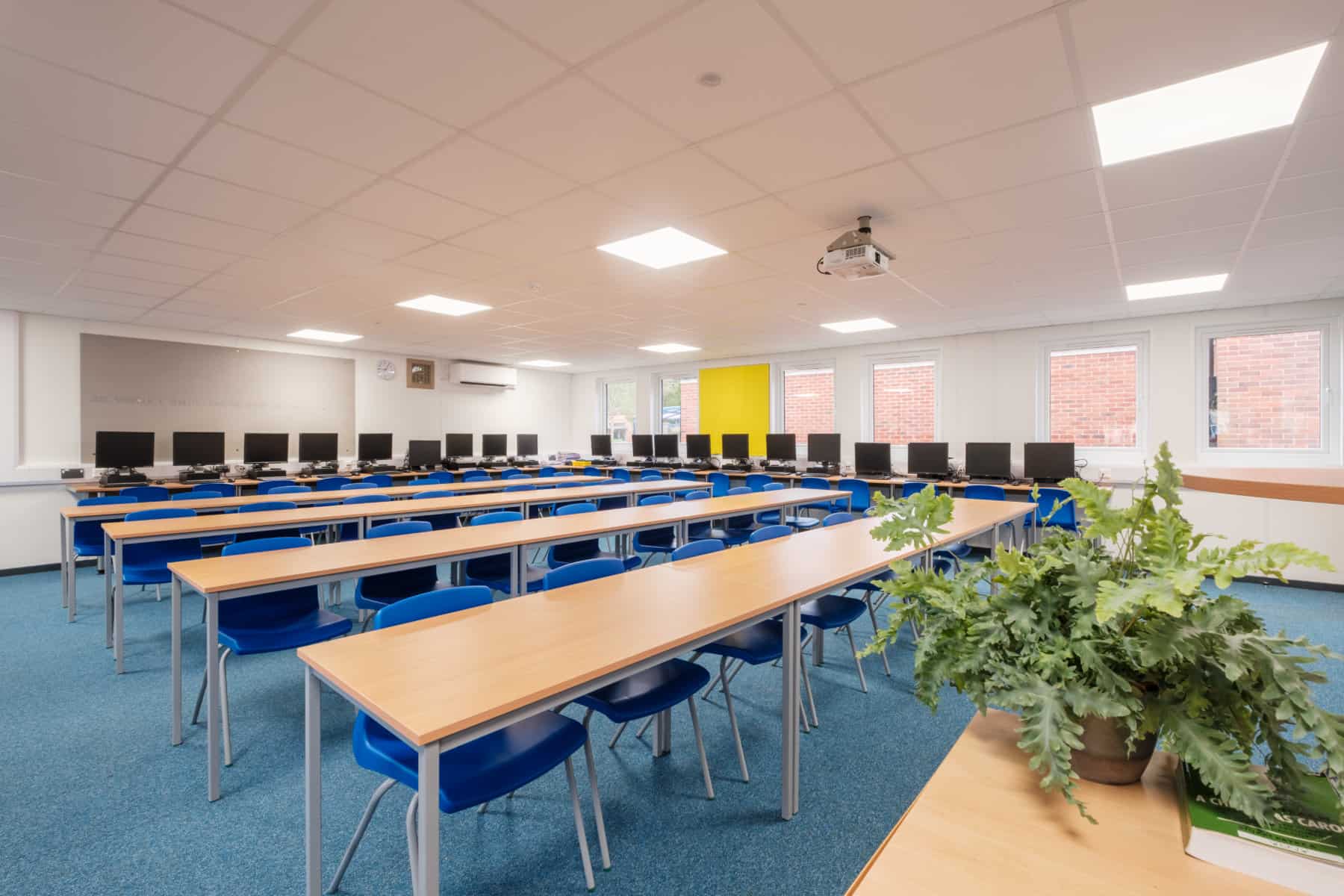 A classroom inside a modular school building. Rows of empty desks and blue chairs are arranged in a computer lab, flanked by workstations with monitors. The environment includes white walls, windows, and a projector. A plant sits on the front desk.