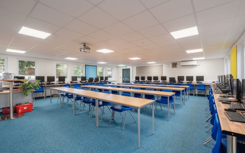 Inside a modular classroom on a refurbished modular building project. A spacious computer lab contains rows of desks with blue chairs, each desk equipped with computers. Bright overhead lights illuminate the room with windows providing natural light; a projector hangs from the ceiling.