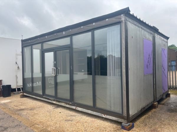 A metal container with large glass front doors and windows stands on a paved area outside, with a cloudy sky overhead. Purple insulation panels are embedded on its sides.