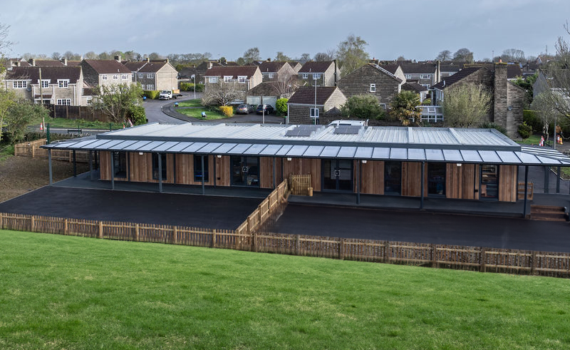 A modern, rectangular building with a metal roof and wooden paneling stands on an asphalt lot surrounded by grass, fencing, and residential houses in a suburban area.