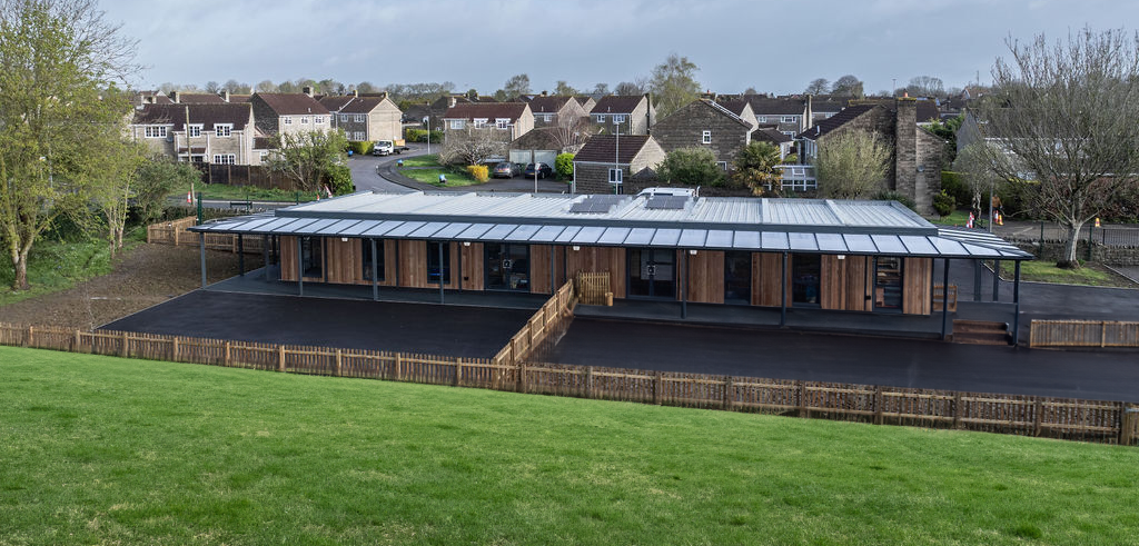 A modern, rectangular building with a metal roof and wooden paneling stands on an asphalt lot surrounded by grass, fencing, and residential houses in a suburban area.