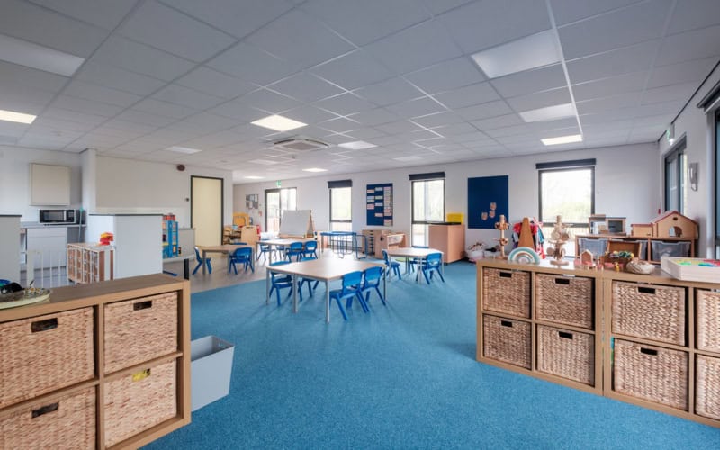 A spacious classroom featuring small tables and blue chairs in an organized layout. Storage units with wicker baskets line the room, with educational materials and toys dispersed throughout the brightly lit space.