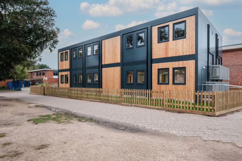 A modern two-story modular building with wood and dark panels, surrounded by a wooden fence, is situated along a paved walkway with trees and another brick structure in the background.