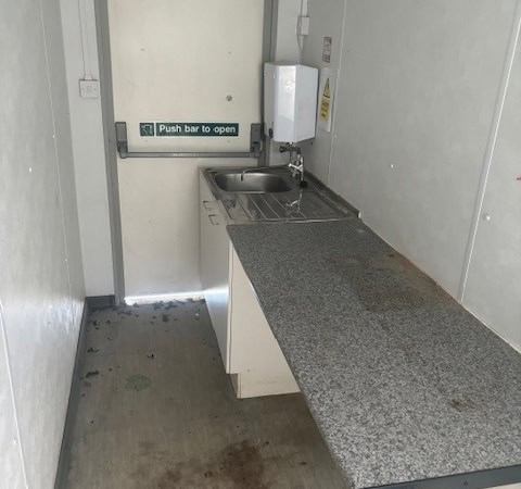 A small, narrow kitchen space with a countertop and sink beside a door labeled "Push bar to open." The area appears unclean with dirt and stains on the floor and countertop.