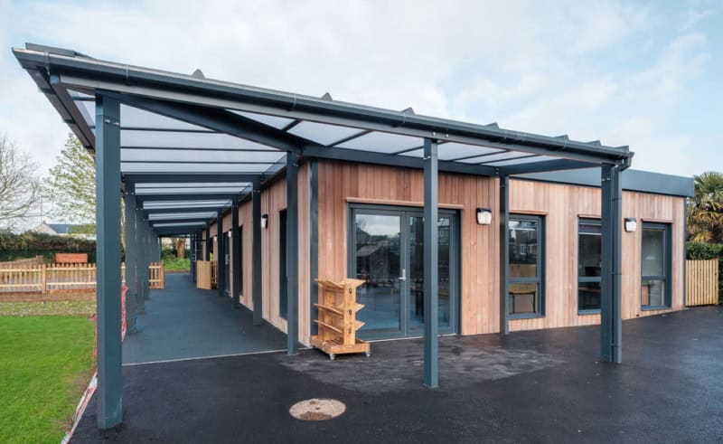 Building with wooden exterior panels has a covered walkway supported by metal pillars; situated in a school yard with grass and wooden fences.