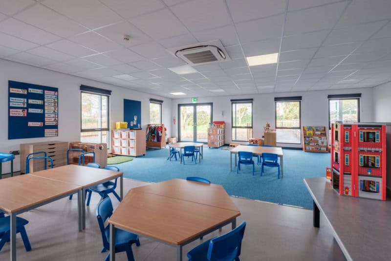 Classroom with blue chairs and tables, bookshelves, and organized bins; walls have educational displays, windows, and a door leading outside, with a carpeted area for activities.