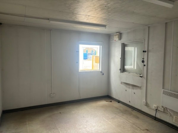 A small, empty room with white walls, a single window with a view of construction, a wall with a windowed counter, and minimal lighting. It appears vacant and undecorated.