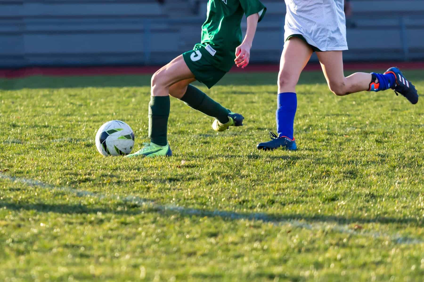 Image shows a field with children playing football. We can only see their legs and they are chasing a football.