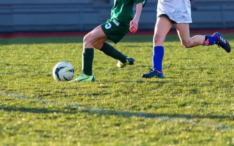 Image shows a field with children playing football. We can only see their legs and they are chasing a football.